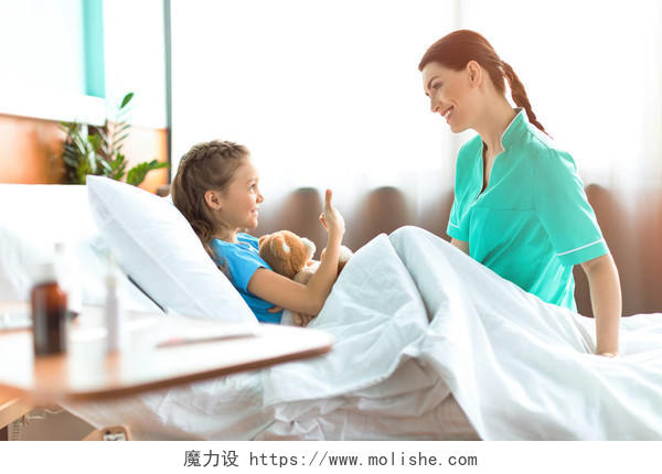 可爱的小女孩躺在医院床上的玩具熊和谈话与微笑的护士